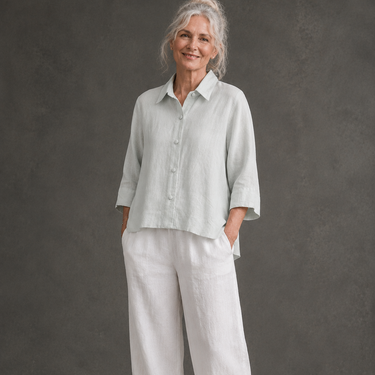 An older woman stands barefoot against a textured gray studio backdrop, wearing a light sage green linen button-up shirt and matching wide-leg linen pants.
