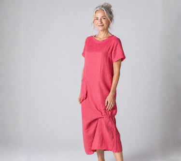 A woman modeling a short-sleeved, loose-fitting midi dress in a vibrant pink fabric against a neutral gray studio background.