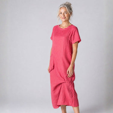 A woman modeling a short-sleeved, loose-fitting midi dress in a vibrant pink fabric against a neutral gray studio background.