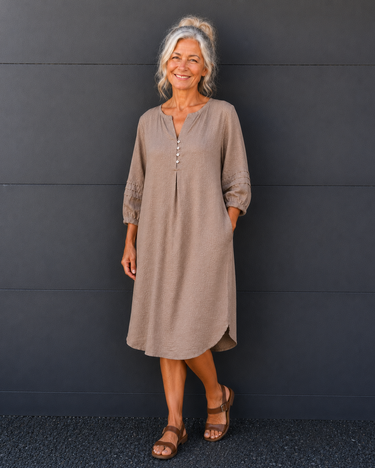 A smiling older woman with gray hair stands against a dark gray paneled wall wearing a loose taupe-brown midi tunic dress with lace sleeve details and brown sandals.