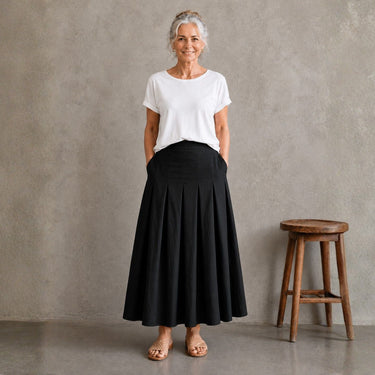 An older woman stands in a studio wearing a white short-sleeve top and a long black pleated skirt, with a wooden stool beside her against a textured gray wall.