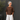 A woman with gray hair in a bun wearing a brown short-sleeve top and white pants, standing against a dark gray wall next to a potted plant.