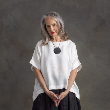An older woman with gray hair stands against a textured gray studio background, wearing a loose white short-sleeve top, a black skirt, and a black pendant necklace.