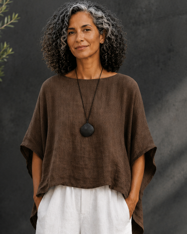 A woman stands outdoors wearing a loose brown short-sleeve top and wide-leg white pants, with a long black pendant necklace and tan sandals. The background features a dark textured wall, gravel, and a potted plant.