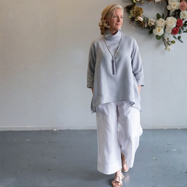 A woman wearing a light gray mock-neck top and white wide-leg pants, standing in a studio with a gray floor and a floral wall hanging in the background.