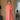A woman wearing a loose, short-sleeved coral pink midi dress with an asymmetrical hem, standing in a modern room with light wood floors and beige curtains.