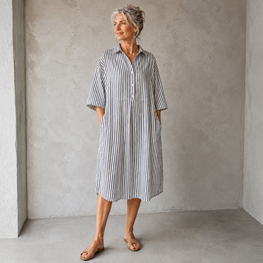 A woman wearing a loose, short-sleeve button-front dress with vertical gray and white stripes, standing in a minimalist gray room.