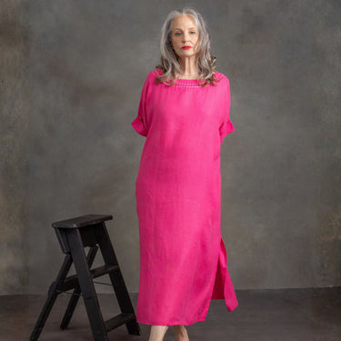 A woman with gray hair wearing a bright pink linen dress with short sleeves and a square neckline, standing next to a small black wooden stool against a textured gray studio backdrop.