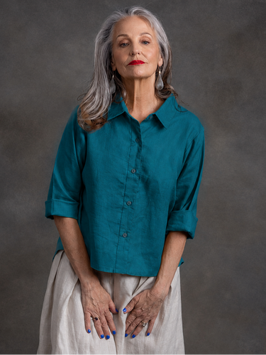 An older woman with gray hair stands against a dark gray textured backdrop, wearing a teal button-up shirt with rolled sleeves and a light beige skirt.