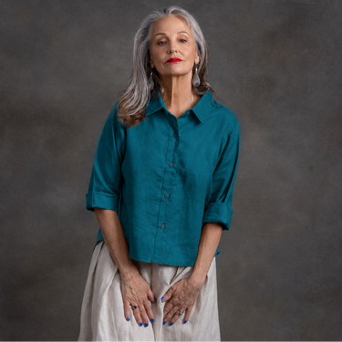 An older woman with gray hair stands against a dark gray textured backdrop, wearing a teal button-up shirt with rolled sleeves and a light beige skirt.