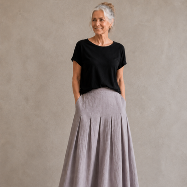 An older woman stands against a textured gray wall wearing a black short-sleeve top and a long, pleated light gray skirt with side pockets, paired with tan sandals.
