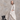 An older woman with gray hair stands barefoot in a studio setting, wearing a long, loose beige linen tunic over white pants. She is posed next to a small black wooden stool against a textured gray plaster background.