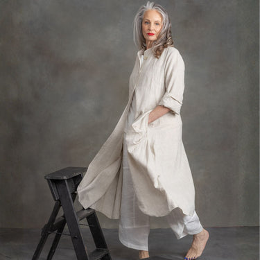 A woman with gray hair stands barefoot in a studio setting, wearing a loose, long-sleeved cream linen tunic with a matching long drape and wide-leg pants. She is posed next to a small black wooden step stool against a textured gray backdrop.