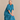 An older woman with gray hair sits on a dark wooden stool, wearing a loose-fitting teal blue dress. The background is a neutral beige textured wall.