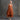 A woman with gray hair and red lipstick wearing a rust-orange linen dress, standing barefoot against a textured gray studio wall.