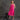 A woman stands barefoot in a studio wearing a bright pink sleeveless dress, with a small black stool beside her against a textured gray backdrop.