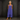 A woman wearing a sleeveless, tiered blue maxi dress with side pockets, standing against a dark textured studio backdrop.