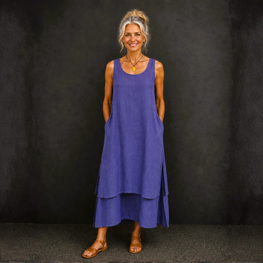 A woman wearing a sleeveless, tiered blue maxi dress with side pockets, standing against a dark textured studio backdrop.