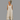 A woman wearing a sleeveless, high-low tiered maxi dress in a light beige linen-like fabric, standing against a solid gray background.
