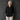 An older woman with gray hair stands against a dark textured studio wall wearing a loose black button-front blouse, light beige wide-leg pants, and black shoes.