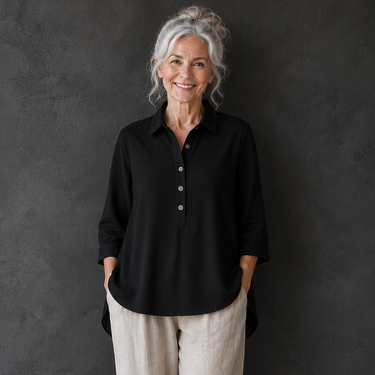 An older woman with gray hair stands against a dark textured studio wall wearing a loose black button-front blouse, light beige wide-leg pants, and black shoes.