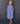 A smiling older woman stands against a dark gray paneled wall wearing a loose, long-sleeve midi dress in a dusty periwinkle blue color. The dress features a split V-neckline with decorative buttons, gathered cuffs, and side pockets.