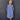 A smiling older woman stands against a dark gray paneled wall wearing a loose, long-sleeve midi dress in a dusty periwinkle blue color. The dress features a split V-neckline with decorative buttons, gathered cuffs, and side pockets.