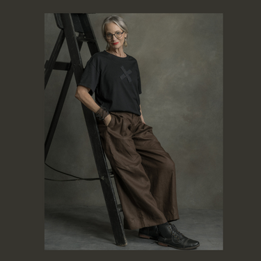 A model wearing cocoa brown wide leg linen trousers with a black top, standing next to a ladder in a studio setting.