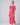 A woman modeling a short-sleeved, loose-fitting midi dress in a vibrant pink fabric against a neutral gray studio background.