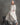 A woman with gray hair stands barefoot in a studio setting, wearing a loose, long-sleeved cream linen tunic with a matching long drape and wide-leg pants. She is posed next to a small black wooden step stool against a textured gray backdrop.