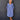 A smiling older woman stands against a dark gray paneled wall wearing a loose, long-sleeve midi dress in a dusty periwinkle blue color. The dress features a split V-neckline with decorative buttons, gathered cuffs, and side pockets.