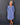A smiling older woman stands against a dark gray paneled wall wearing a loose, long-sleeve midi dress in a dusty periwinkle blue color. The dress features a split V-neckline with decorative buttons, gathered cuffs, and side pockets.