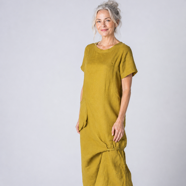 A woman modeling a short-sleeved, midi-length linen dress in a mustard yellow color, standing against a light gray studio background.