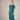 A woman modeling a loose-fitting, short-sleeved midi dress in a deep teal green color, standing against a textured beige studio backdrop.