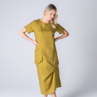 A woman modeling a short-sleeved olive yellow linen midi dress with a gathered front and large patch pockets, set against a plain light gray studio background.