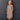 A smiling older woman with gray hair stands against a dark gray paneled wall wearing a loose taupe-brown midi tunic dress with lace sleeve details and brown sandals.