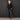 An older woman stands against a dark textured wall wearing a black button-front collared shirt with three-quarter sleeves and dark charcoal wide-leg pants, paired with tan flat sandals.