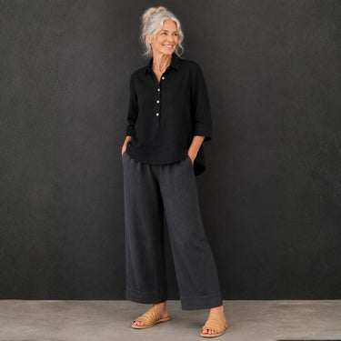 An older woman stands against a dark textured wall wearing a black button-front collared shirt with three-quarter sleeves and dark charcoal wide-leg pants, paired with tan flat sandals.