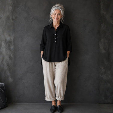 An older woman with gray hair stands against a dark textured wall wearing a black button-front blouse, loose light beige wide-leg pants, and black loafers.