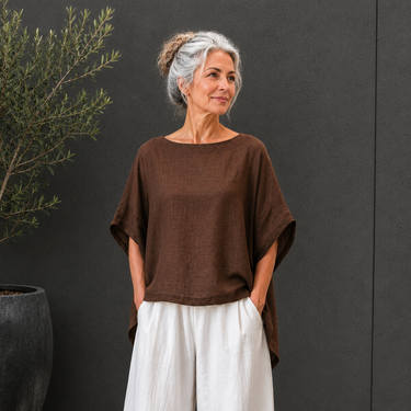 A woman with gray hair in a bun wearing a brown short-sleeve top and white pants, standing against a dark gray wall next to a potted plant.