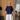 A woman wearing a loose navy blue short-sleeve top and white wide-leg pants, standing in a home interior with beige curtains and light wood flooring.