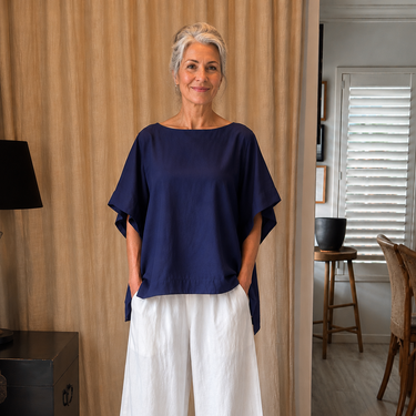 A woman wearing a loose navy blue short-sleeve top and white wide-leg pants, standing in a home interior with beige curtains and light wood flooring.