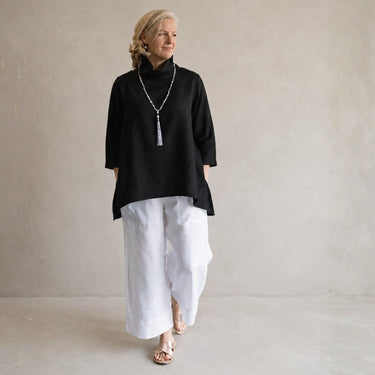 A woman wearing a black turtleneck top with three-quarter sleeves, white wide-leg pants, and a long silver tassel necklace, standing against a textured light gray wall.