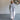 A woman wearing a light gray mock-neck top and white wide-leg pants, standing in a studio with a gray floor and a floral wall hanging in the background.