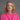 A woman with blonde shoulder-length hair wearing a bright pink top with white embroidered trim, photographed against a neutral gray studio background.