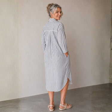 A woman wearing a blue and white vertically striped midi shirt dress, photographed from behind in a minimalist room with a beige wall and concrete floor.