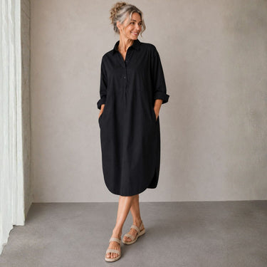 A woman wearing a black long-sleeve button-up shirt dress with rolled sleeves and side pockets, standing in a minimalist studio with a textured gray wall and concrete floor.