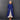 An older woman stands against a dark gray tiled wall wearing a long, vibrant royal blue linen-blend coat over a cream-colored dress.