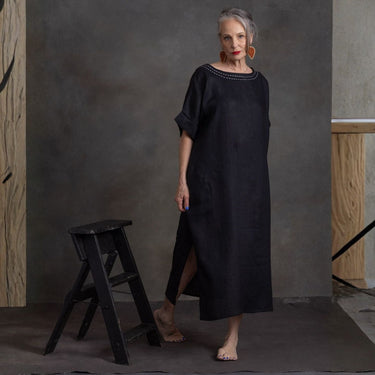 A woman stands barefoot in a studio wearing a loose black midi dress with short sleeves and a decorative embroidered neckline. A small black stool sits beside her against a textured gray backdrop.