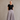 An older woman stands against a textured gray wall wearing a black short-sleeve top and a long, pleated light gray skirt with side pockets, paired with tan sandals.